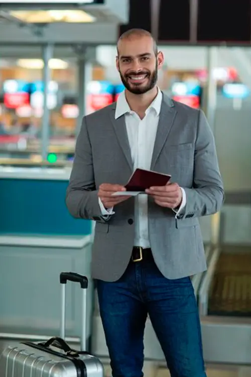 Man in gray blazer and jeans holding passport, standing with suitcase at airport.